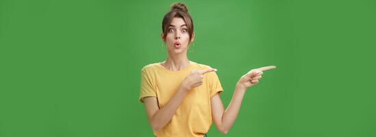 Woman reacting to awesome sales in near shop pointing left and gazing impressed and excited at camera folding lips from desire and amazement staring at camera thrilled posing over green background photo