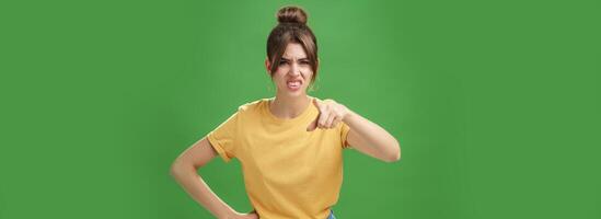 Portrait of arrogant impolite young woman in yellow t-shirt pointing at camera squinting and grimacing from disdain and scorn standing unimpressed judging against green background photo