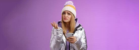 Questioned complicated cute blond girlfriend receive strange message look perplexed confused raising hand shrugging lift eyebrow cannot understand meaning holding smartphone, purple background photo