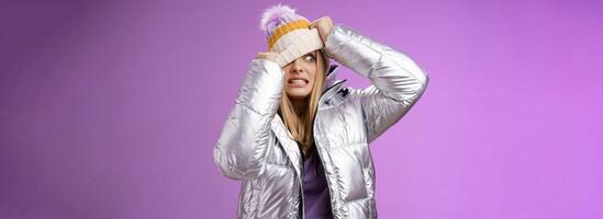 Awkward embarrassed cute blond girl worried pulling hat face hiding from ex-boyfriend pretend not see him standing anxiously clenching teeth peeking one eye left nervous, purple background photo