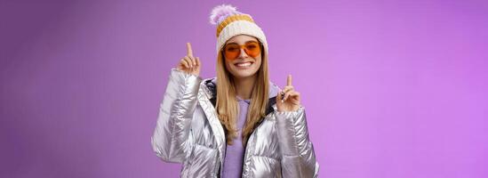Joyful energized entertained cute blond woman having fun enjoy vacation snowy mountain trip wearing sunglasses silver jacket winter hat dancing pointing up amused standing purple background photo