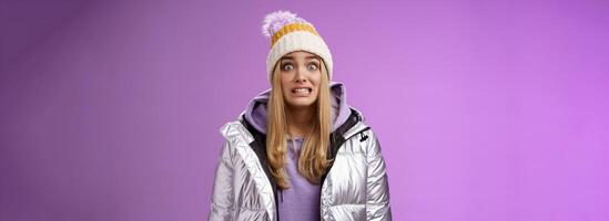 Awkward worried cute timid blond girl in silver jacket hoodie winter hat clench teeth popping eyes camera ooops make mistake standing nervous someone notice, purple background photo