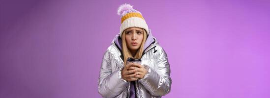 Trembling uncomfortable cute pouting young blond girl feel freezing cold winter snowy weather outside shaking low temperature hold take-away hot coffee cup warming up, purple background photo