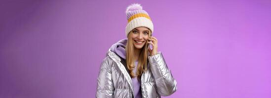 Joyful modern stylish millennial young blond girl talking friends smartphone calling mom from winter resort vacation abroad wearing hat silver jacket hold mobile phone near ear smiling photo