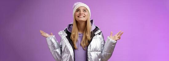 Thankful happy cute attractive blond young 25s woman in winter hat silver trendy jacket raise hands look up grateful god dream come true smiling delighted fulfilled wish, purple background photo