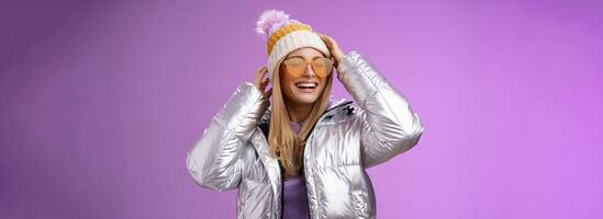 Carefree amused charming blond girlfriend having fun enjoying awesome sunny winter day ski resort vacation wearing sunglasses silver stylish jacket put on hat grinning joyfully, purple background photo