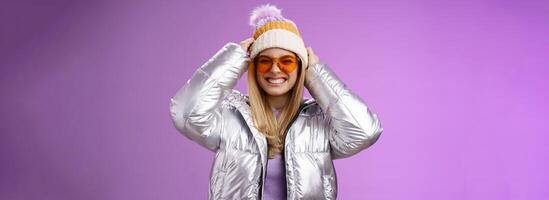 Energized daring sassy young attractive woman having fun friends winter trip learn snowboarding smiling cheeky enjoying vacation put-on hat wearing silver warm jacket sunglasses, purple background photo