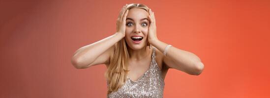 Amused surprised excited young blond attractive woman in silver glittering stylish dress hold head hands widen eyes speechless look camera unbelievable thrill fan see famous person, red background photo