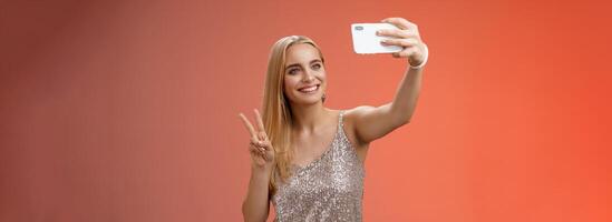 Attractive feminine tender young blond girl 25s in silver stylish dress taking selfie extend arm up show peace gesture smartphone display record video greeting internet fans, standing red background photo