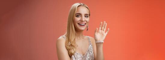 Waist-up studio shot friendly attractive elegant tender blond woman say hi waving raised hand greeting welcoming friend smiling delighted introducing herself hello gesture, red background photo