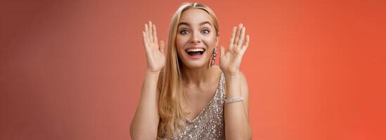Impressed charming entertained blond european 25s woman in stylish silver glamour dress having fun playing peekaboo smiling joyfully hold palms near face laugh amused, standing red background photo