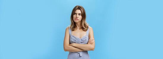 Studio shot of woman thinking seriously being suspicious and hesitant pursing lips holding hands crossed on chest looking at upper right corner with frown, thinking over blue background photo