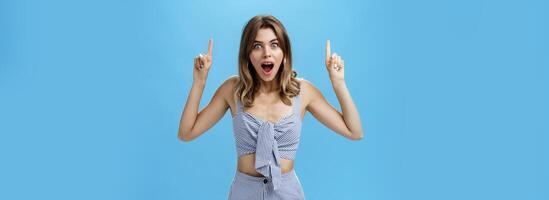 Woman getting surprised dropping jaw from amazement smiling broadly staring at camera astonished raising hands and pointing up at awesome copy space posing against blue background photo