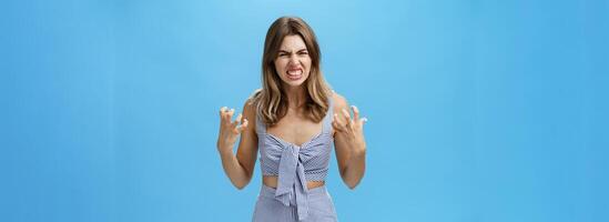 Indoor shot of irritated moody woman having mental breakdown feeling annoyed and pressured squeezing hands in fists from anger wrinkling nose in hate standing hateful over blue background photo