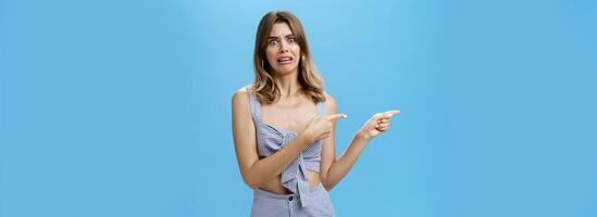 Studio shot of awkward displeased woman pointing left with index fingers making disgusted and dissatisfied expression grimacing from aversion seeing something creepy and awful over blue background photo