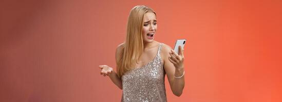 Troubled concerned arrogant young blond woman complaining yelling smartphone cannot call friend no signal holding smartphone look mobile display pissed moody arguging, red background photo