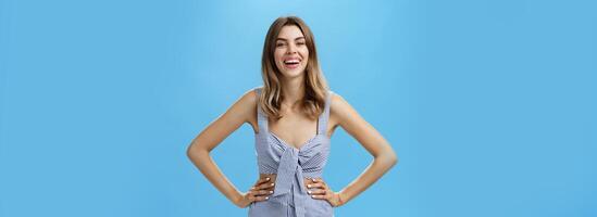 Amused and delighted cute adult female with chestnut hair in casual clothes holding hands on hip laughing and smiling broadly feeling excited during conversation posing against blue background photo