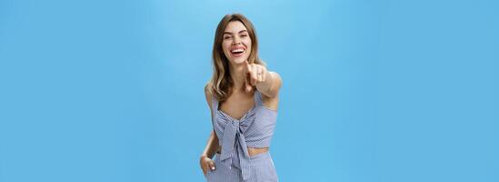 Haha your are funny. Portrait of amused carefree attractive young female in matching outfit pointing at camera as if having fun over photographer laughing out loud being in good mood over blue wall photo