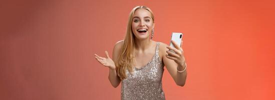 Astonished happy charming blond girl in silver glittering stylish dress holding smartphone amazed liking awesome result edit photo app smiling wondered amused, standing red background