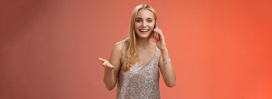 Joyful talkative outgoing attractive blond woman talking friend smartphone gesturing amused smiling broadly retelling fresh rumors after party wearing silver stylish dress, red background photo