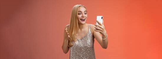 Charming elegant nice blond girl in silver dress talking video call speaking looking smartphone display amused surprised smiling happily have conversation sibling showing prom outfit photo