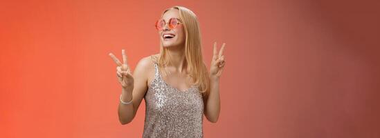 Positive charming european carefree stylish girlfriend having fun dance-floor show victory peace signs look up happily smile in sunglasses glamour glittering dress enjoy celebration, red background photo