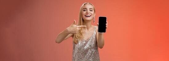 Boastful proud charming elegant blond woman in stylish evening dress show smartphone display proudly pointing mobile phone screen smiling showing photo boyfriend, standing red background