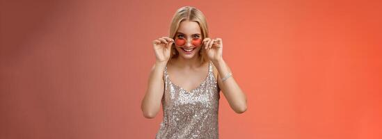 Lucky attractive stylish european blond female having fun friends nightclub wearing silver glittering dress sunglasses look amused curious peek camera intrigued, standing red background photo