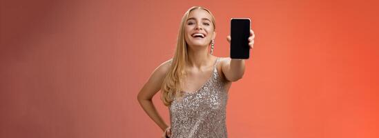 Proud joyful charming cheerful blond european woman in stylish silver shiny dress hold hand waist confident extend arm showing smartphone display present awesome new app device, red background photo