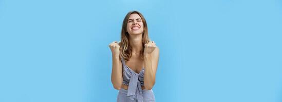 Girl triumphing raising hands in cheer from happiness and victory standing pleased and excited with closed eyes and raised head celebrating successful deal posing delighted against blue background photo