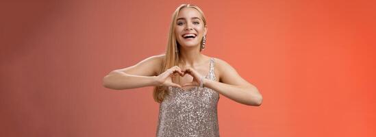 Adorable happy charming lovely blond european girl in silver dress express love positivity show heart gesture confess sympathy passionate feelings towards boyfriend smiling broadly, red background photo