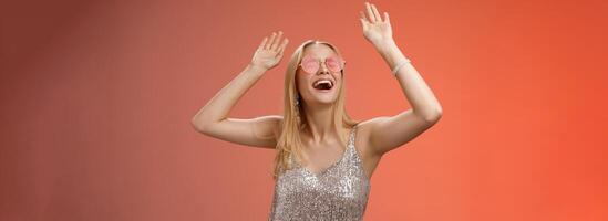 Excited chilling energized young blond woman in silver stylish glittering dress sunglasses raise hands up having fun dancing dance-floor nightclub throw party celebrate b-day, red background photo