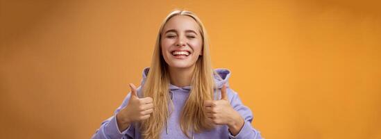Close-up carefree delighted positive lucky young blond woman in hoodie close eyes joyfully celebrating good result show thumbs-up gesture agree approving awesome idea, orange background photo