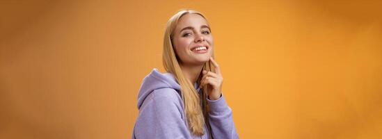 Attractive sassy self-assured caucasian blond female in purple hoodie turning camera touching chin cheeky smile camera check out interesting thing gazing forward intrigued, standing orane background photo