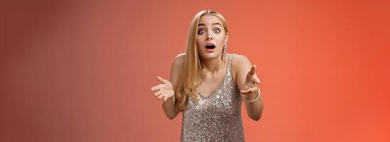 Impressed overwhelmed blond woman retelling exciting story gesturing astonished widen eyes shrugging explaining what happened talking passionately in silver glamour dress standing red background photo