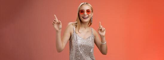 Stylish carefree fabulous young blond woman in silver dress red sunglasses having fun dancing joyfully dance-floor nightclub smiling broadly enjoying spend time friends, standing studio background photo