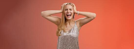 Upset distressed pressured fed up panicking young blond woman in dress scream cry heart out being heartbroken close eyes hold hands head mentaly unstable standing sad depressed red background photo