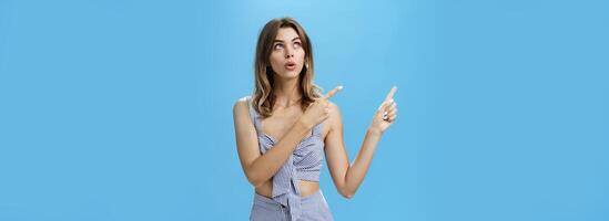 Woman getting interested in curious thing open mouth looking and pointing at upper left corner with intrigued focused expression being fascinated and surprised posing against blue background photo