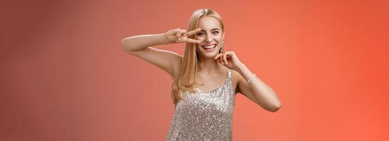 Charismatic funny amused charming blond girl 25s having fun dancing red background in silver trendy glittering party dress show peace disco gesture near eye smiling laughing carefree photo