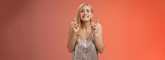 Nervous hopeful blond young woman in silver trendy dress close eyes grimacing eager wish come true cross fingers good luck praying supplicating anticiapting fortune, standing worried red background photo