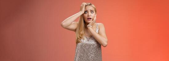 Shocked frightened insecure panicking woman blond hairstyle in silver dress touch head pop eyes afraid scared seing terrifying crime standing stupor speechless gasping shook, red background photo