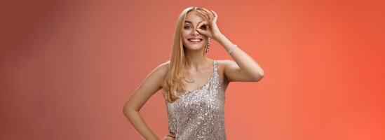 Charming elegant blond european carefree woman in silver glittering evening dress show no problem okay sign look through eye smiling delighted check out awesome promo intrigued, red background photo