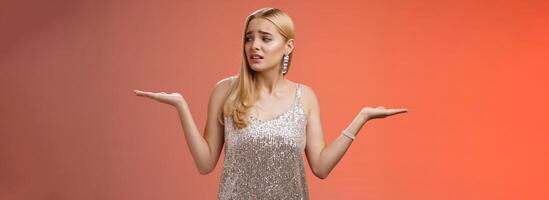 Anxious insecure young cute blond girl feel lost unsure shrugging hands sideways weighing decisions look right seek help standing lost nervous hesitating red background in silver glittering dress photo