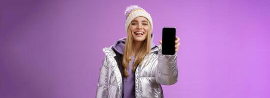 Sassy delighted cheerful blond woman recommend use app edit perfect pictures holding smartphone showing mobile phone display proudly satisfactory smile camera, standing purple background photo