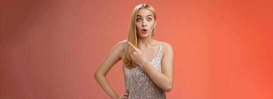 Wondered amused attractive blond woman talking party widen eyes say wow pointing questioned right direction see famous person standing surprised impressed gaze camera, red background photo