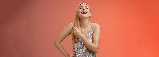 Fabulous carefree attractive blond woman in silver evening party dress laughing out loud have fun raise hand joyfully up pointing right index finger enjoying awesome sense humour, red background photo