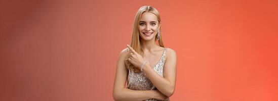 Confident attractive feminine glamour blond young woman in silver glittering dress smiling broadly pointing up discuss recent news promo sales standing friendly grinning camera red background photo