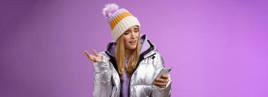 Upset disappointed attractive whining gloomy blond girl in silver jacket standing outside hat holding smartphone shrugging raising hand dismay complaining slow mobile internet, purple background photo