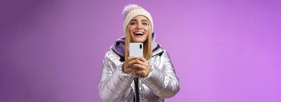 Amused excited attractive blond girlfriend holding smartphone up recording video boyfriend step snowboard first time capturing memories mobile camera standing happily purple background photo