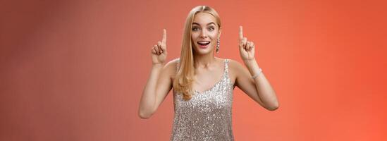 Charming amazed blond european woman in fabulous silver glittering dress raise hands point up amused enjoying watching shooting stars, fireworks gaze camera excited happy surprised, red background photo